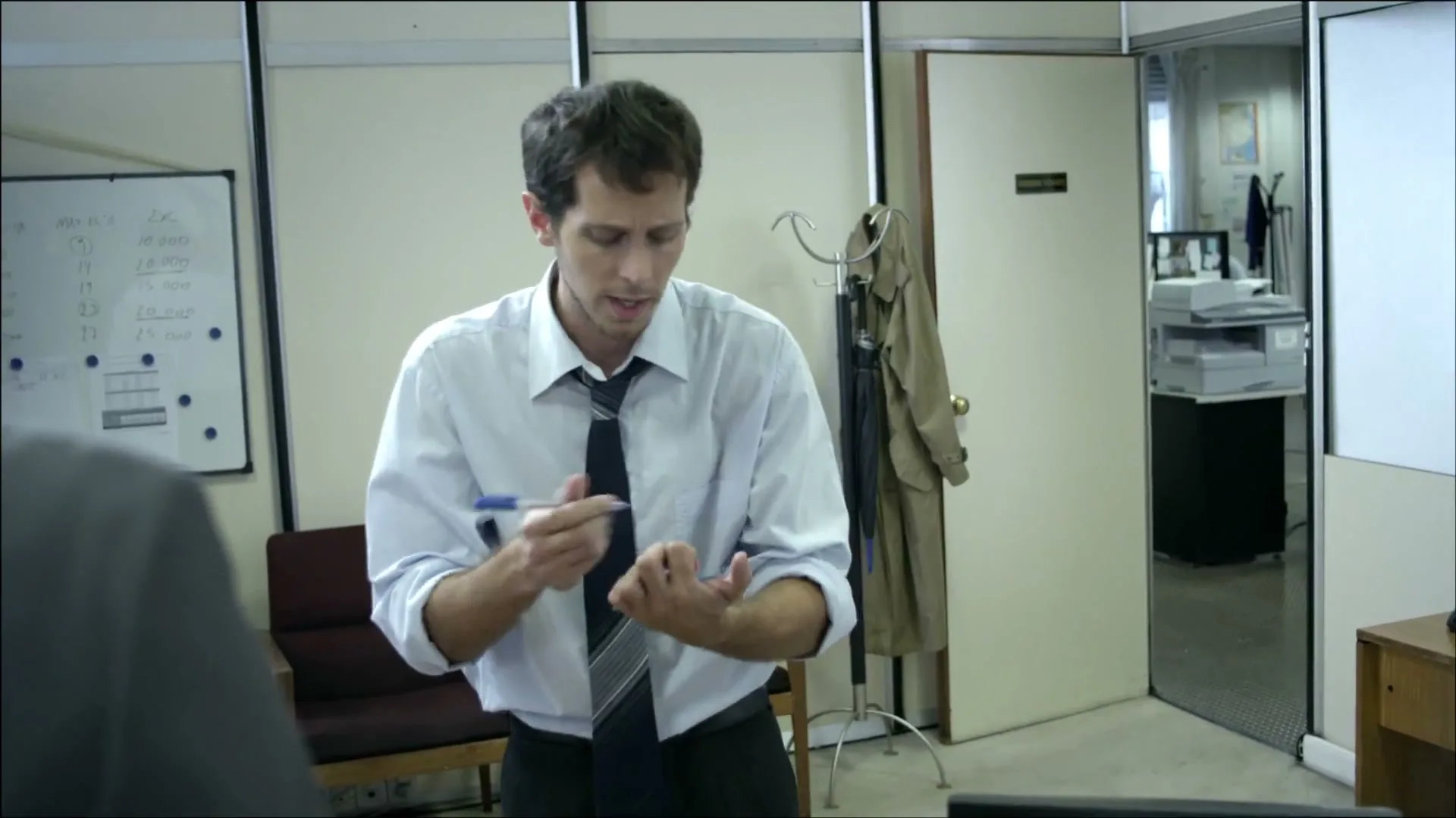 A man in a white shirt and tie, looking stressed, writes on his hand with a blue pen in a brightly lit office.