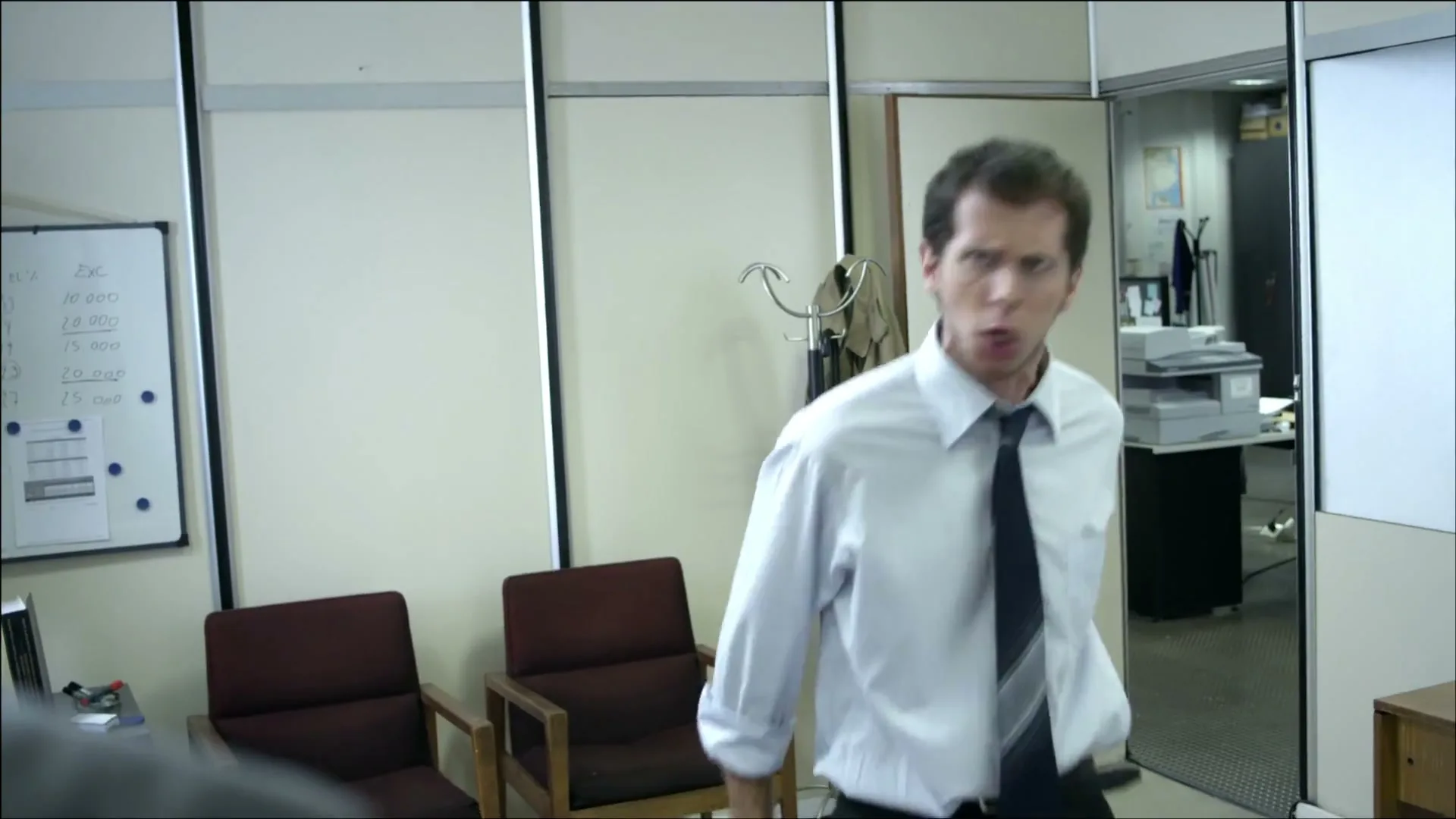 A man in a white shirt and dark tie yells at another person, whose back is to the camera, in a brightly lit office.