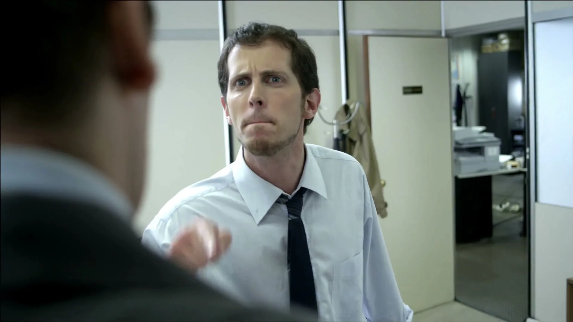 A man in a white shirt and tie argues with another person, pointing his finger in an office setting.