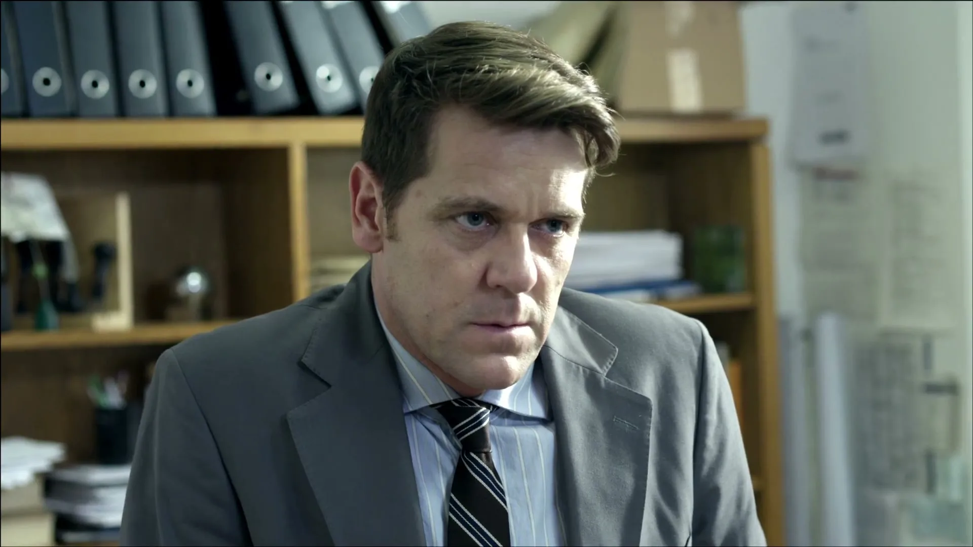 A man in his 40s, wearing a suit and tie, looks intently at someone off-camera in an office setting.