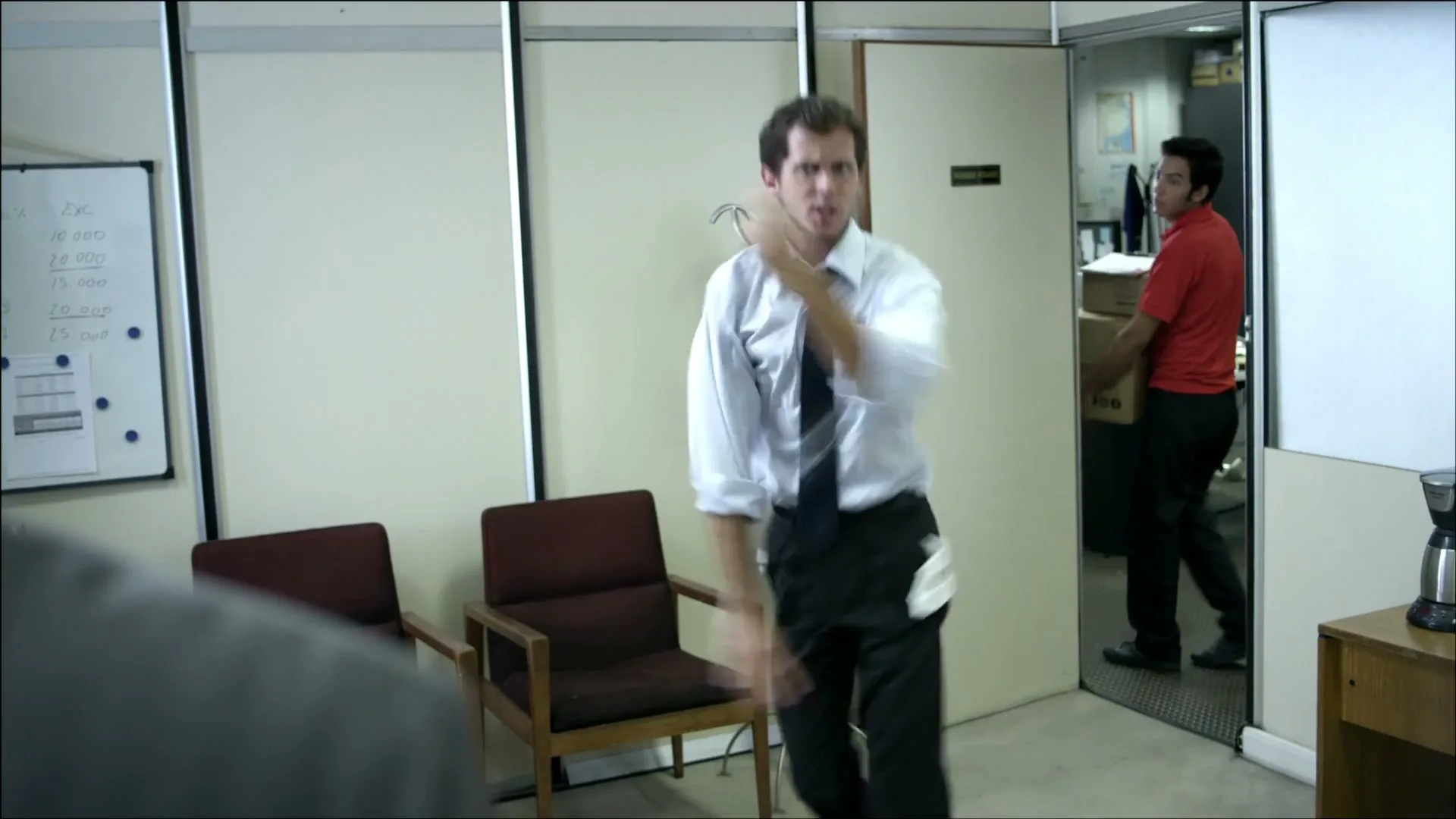 A man in a white shirt and tie dances wildly in an office, while another man in a red shirt carries boxes in the background.