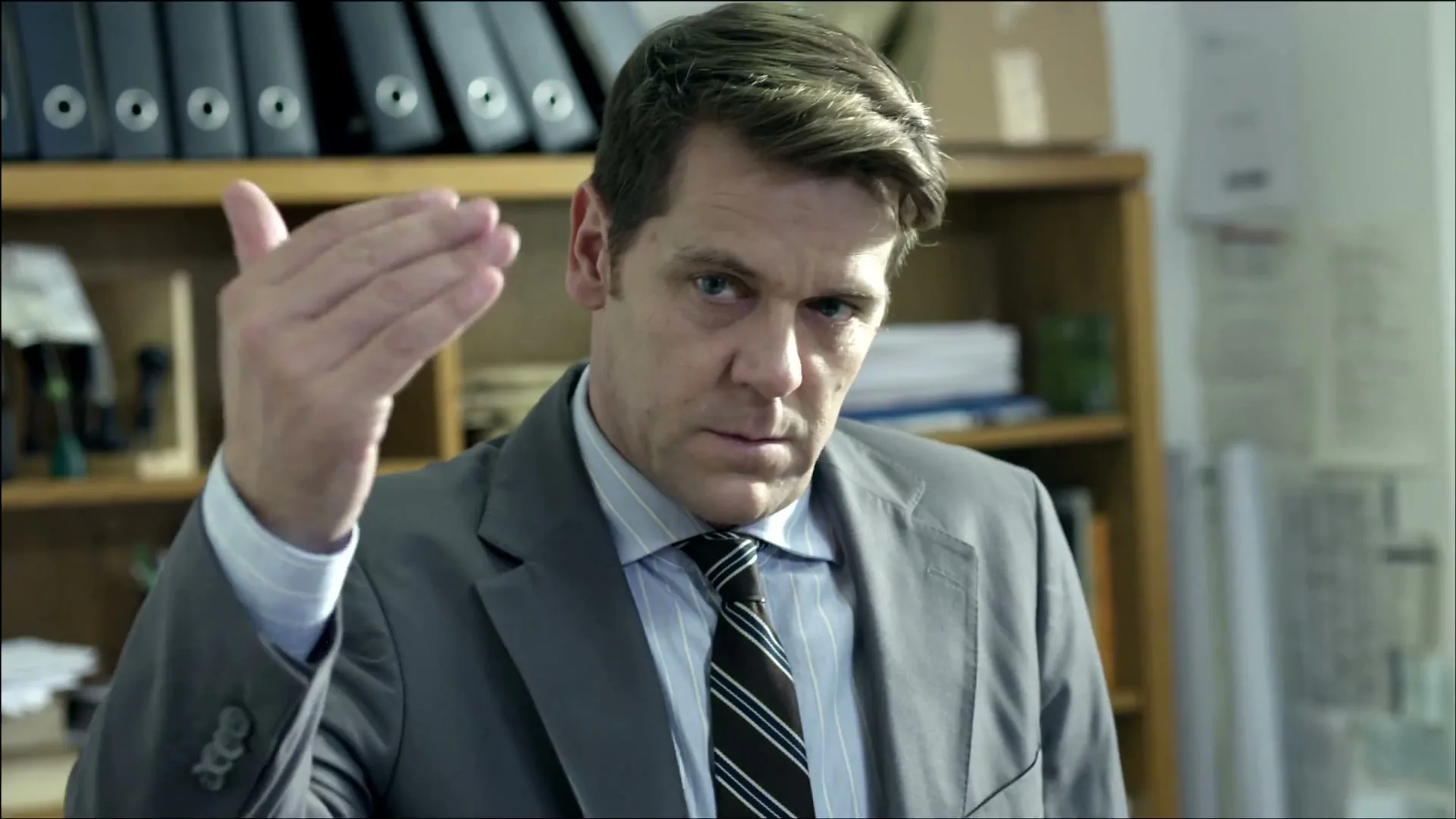 A man in a suit and tie sits at a desk in an office, looking serious and then making a gesture with his hand.