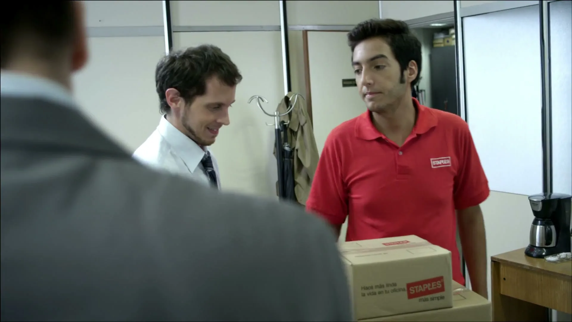 A man in a white shirt and tie dances excitedly while a delivery man in a red Staples shirt delivers boxes to an office worker in an office setting.