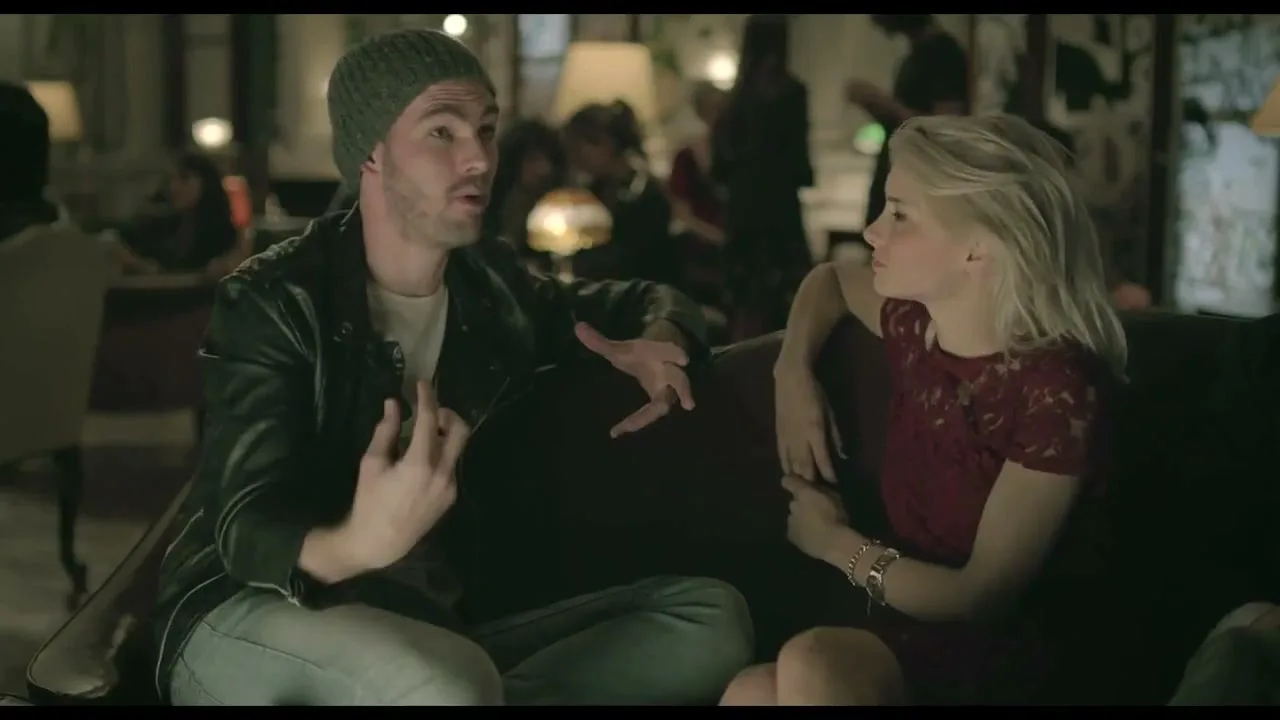 A man in a beanie and leather jacket talks animatedly to a blonde woman in a red dress, both sitting on a couch in a dimly lit, crowded room.