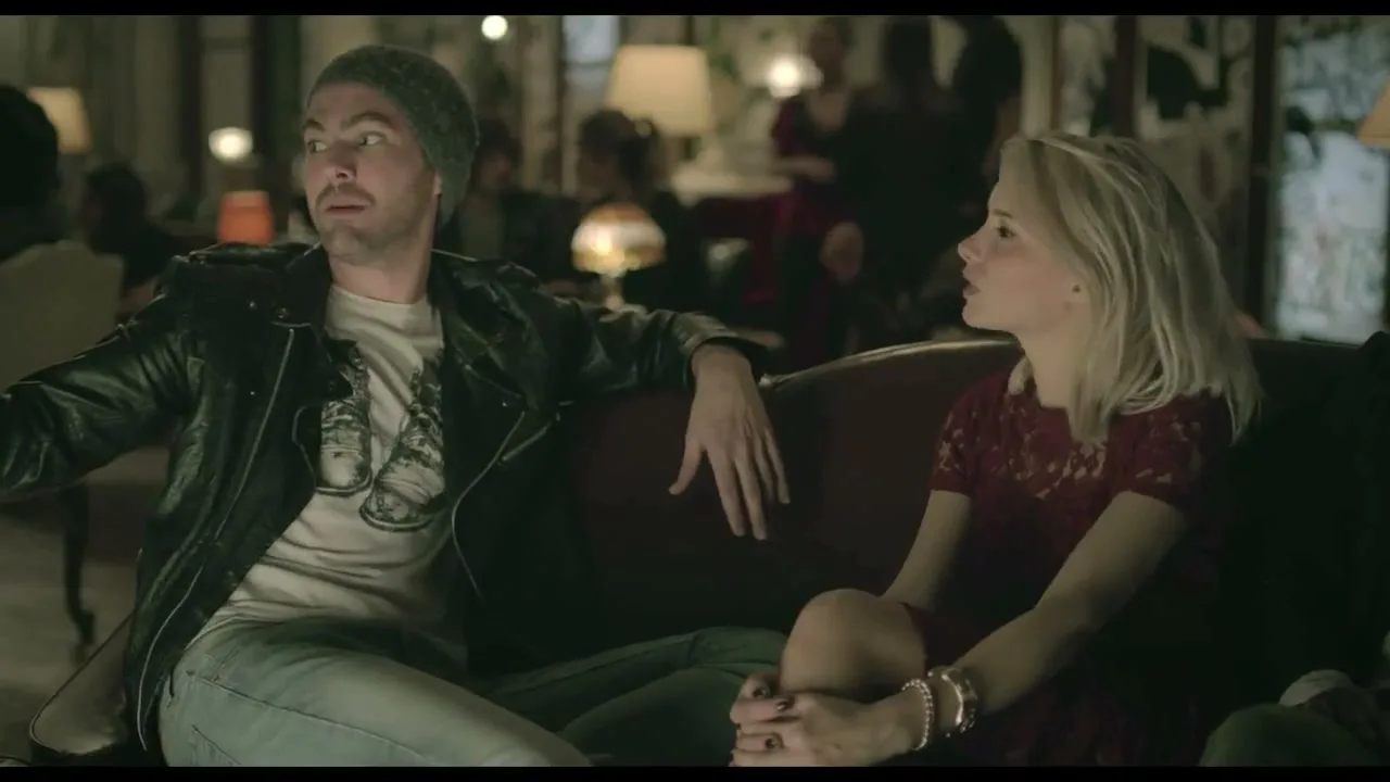 A young man in a beanie and leather jacket talks animatedly to a young woman in a red dress, sitting on a couch in a dimly lit, crowded room.