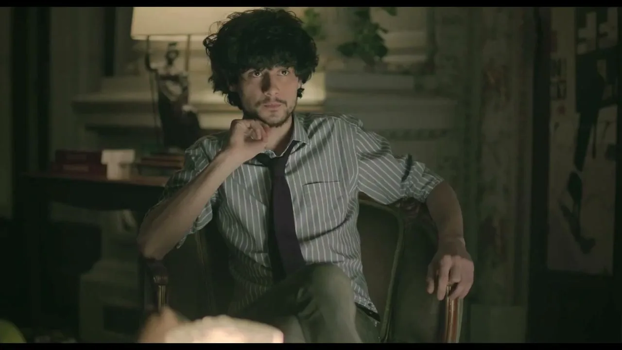 A young man with curly hair and a beard sits in a chair, looking thoughtful in a dimly lit room.