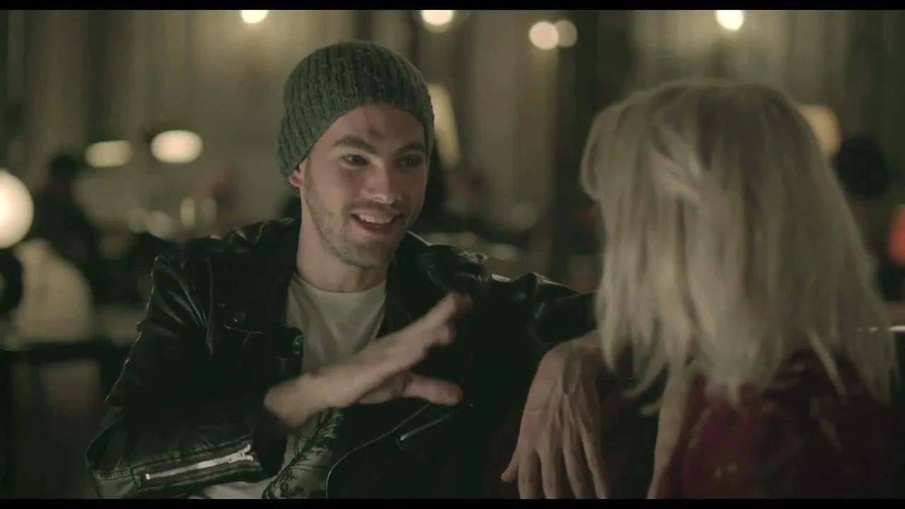 A young man in a beanie and leather jacket talks animatedly to a blonde woman in a dimly lit bar or restaurant.