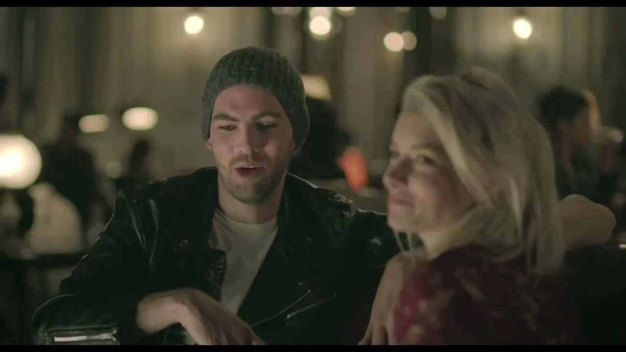 A man in a beanie and leather jacket talks to a blonde woman in a dimly lit bar, gesturing with his hand.