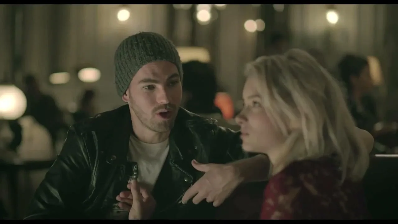 A man in a beanie and leather jacket talks animatedly to a woman with blonde hair in a dimly lit restaurant or bar.