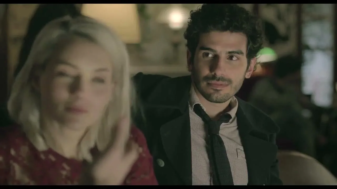 A blonde woman in a red lace top talks to a dark-haired man in a coat and tie, both sitting in a dimly lit interior.