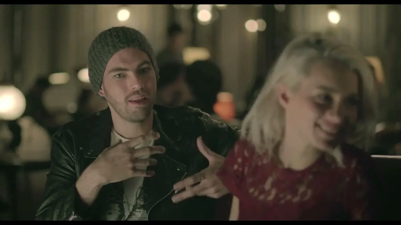 A young man in a beanie and leather jacket talks animatedly to a young woman with blonde hair in a dimly lit bar.