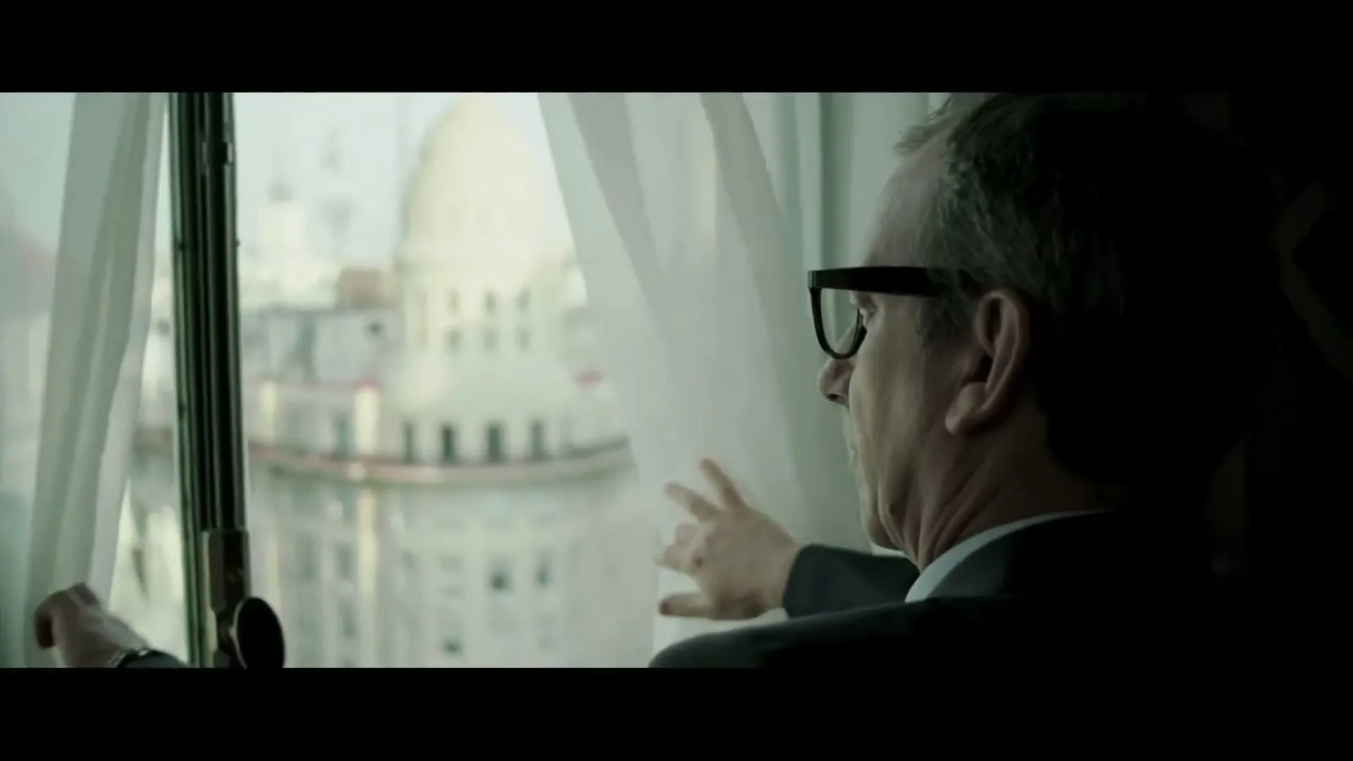 A man in his 50s, wearing a suit and glasses, looks out a window at a city skyline with a large domed building.