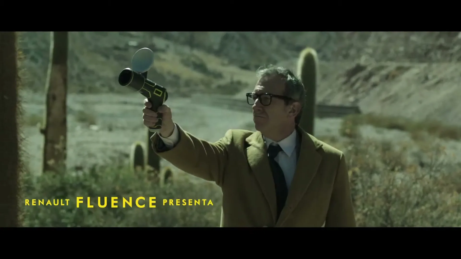 A man in a suit and glasses holds a device like a gun, looking into the distance in a desert landscape with cacti.