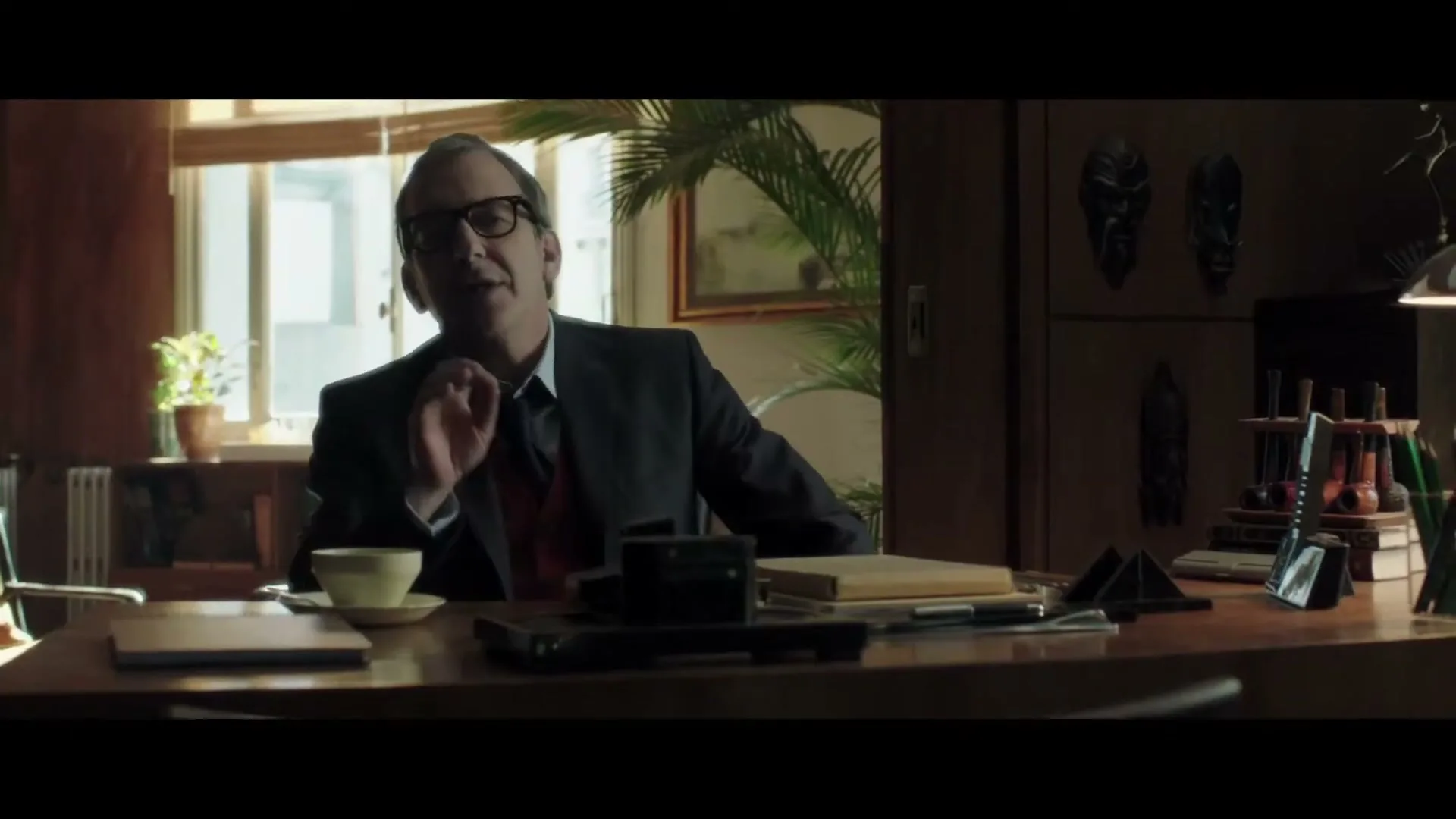 A middle-aged man in a suit and glasses sits at a desk in a dimly lit office, talking and gesturing with his hand.