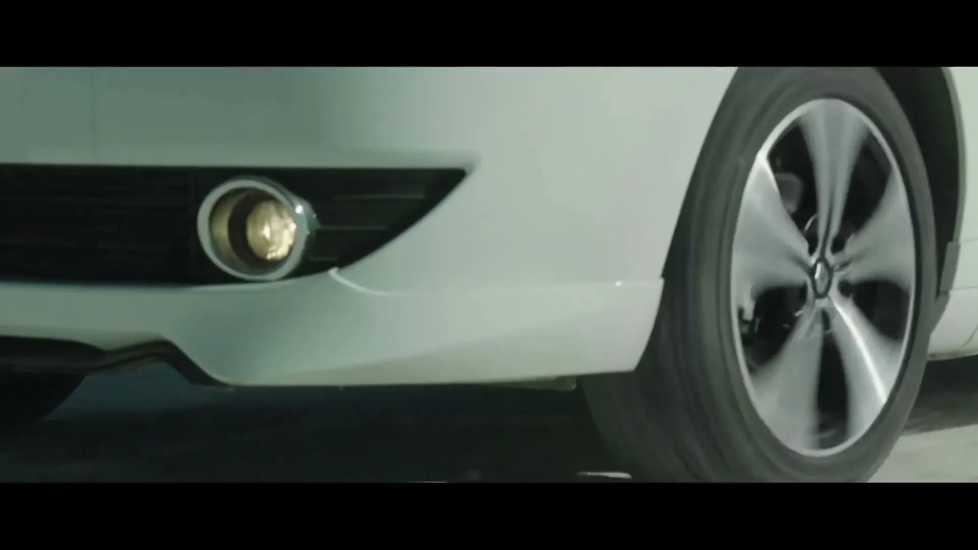 A white car's front bumper and spinning wheel are shown in a medium shot, highlighting its movement on a road.