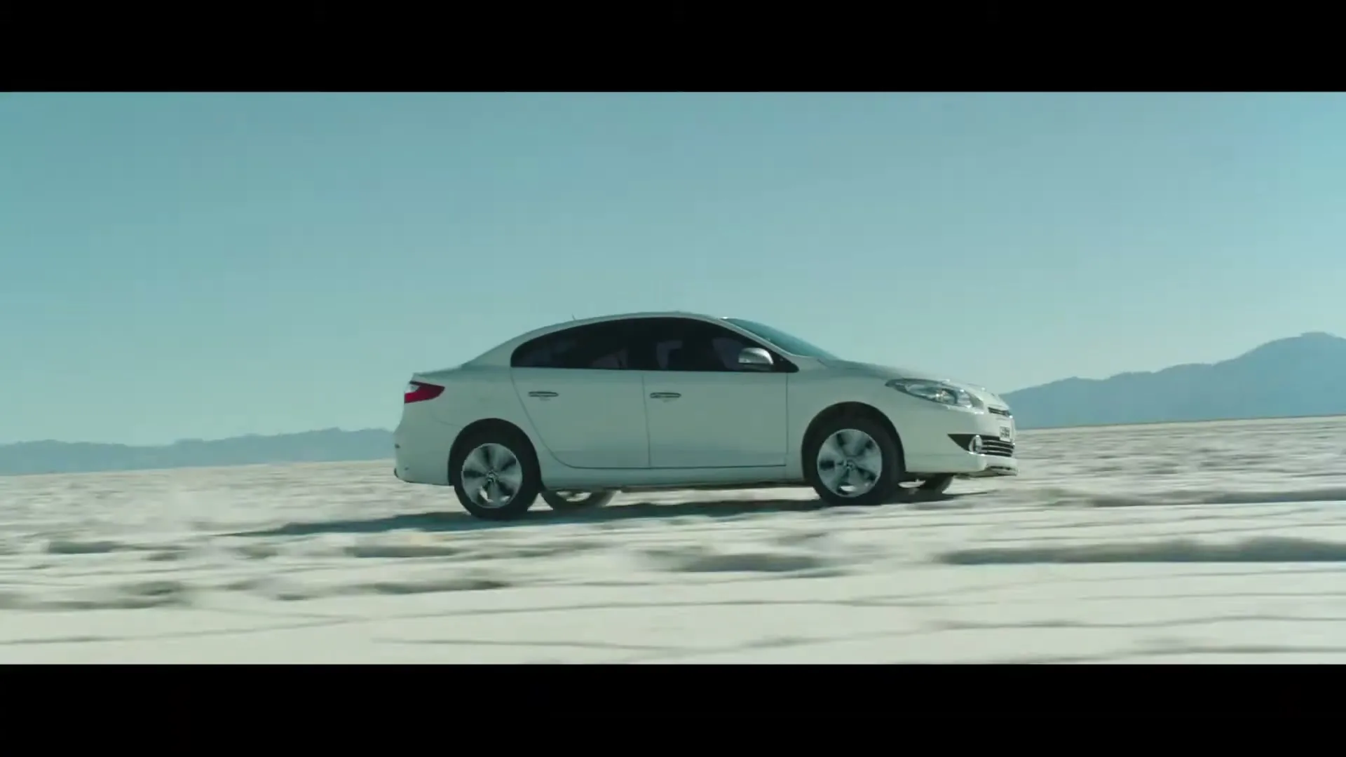 A white sedan drives across a vast, flat, white salt desert under a clear blue sky, with distant mountains on the horizon.