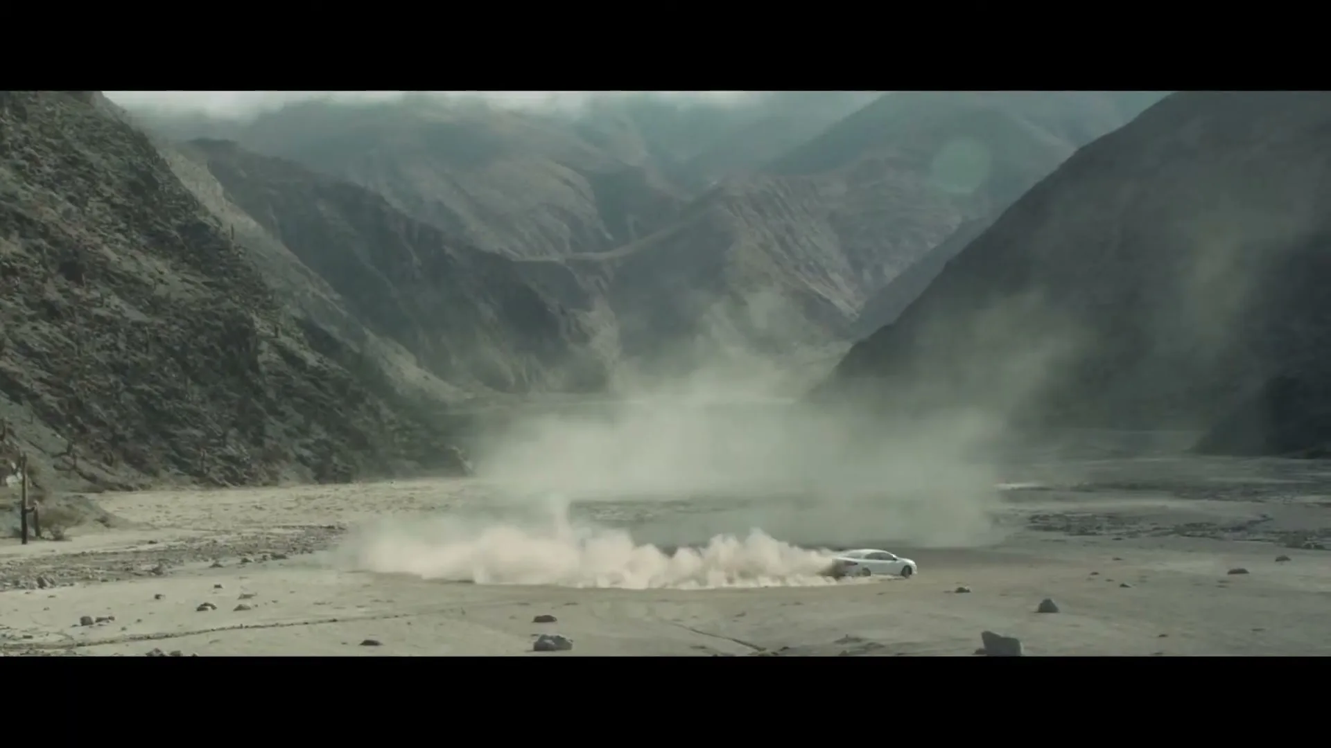 A white car drifts in a dry riverbed, kicking up a large cloud of dust, surrounded by towering mountains under a cloudy sky.