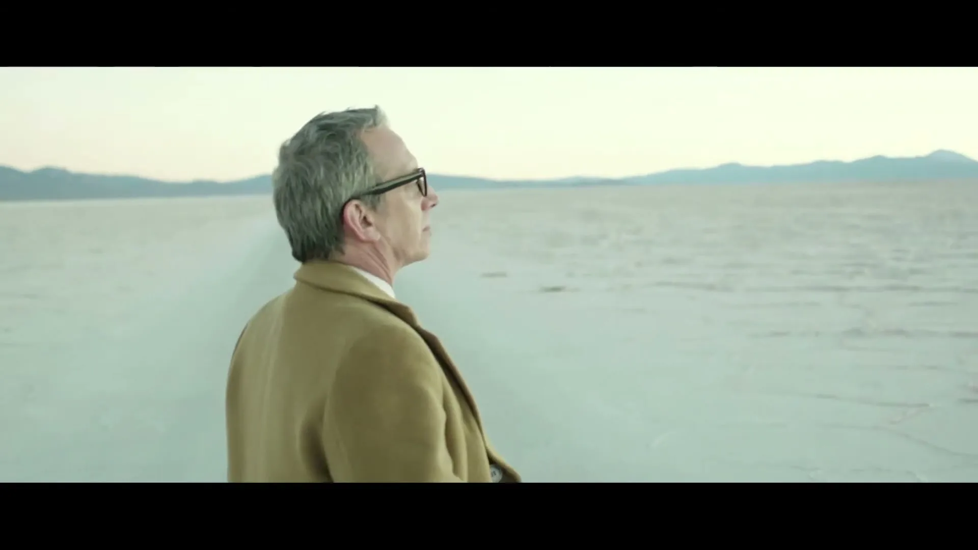 A man with gray hair and glasses, wearing a brown coat, stands in a vast, desolate salt flat, looking out at the horizon.