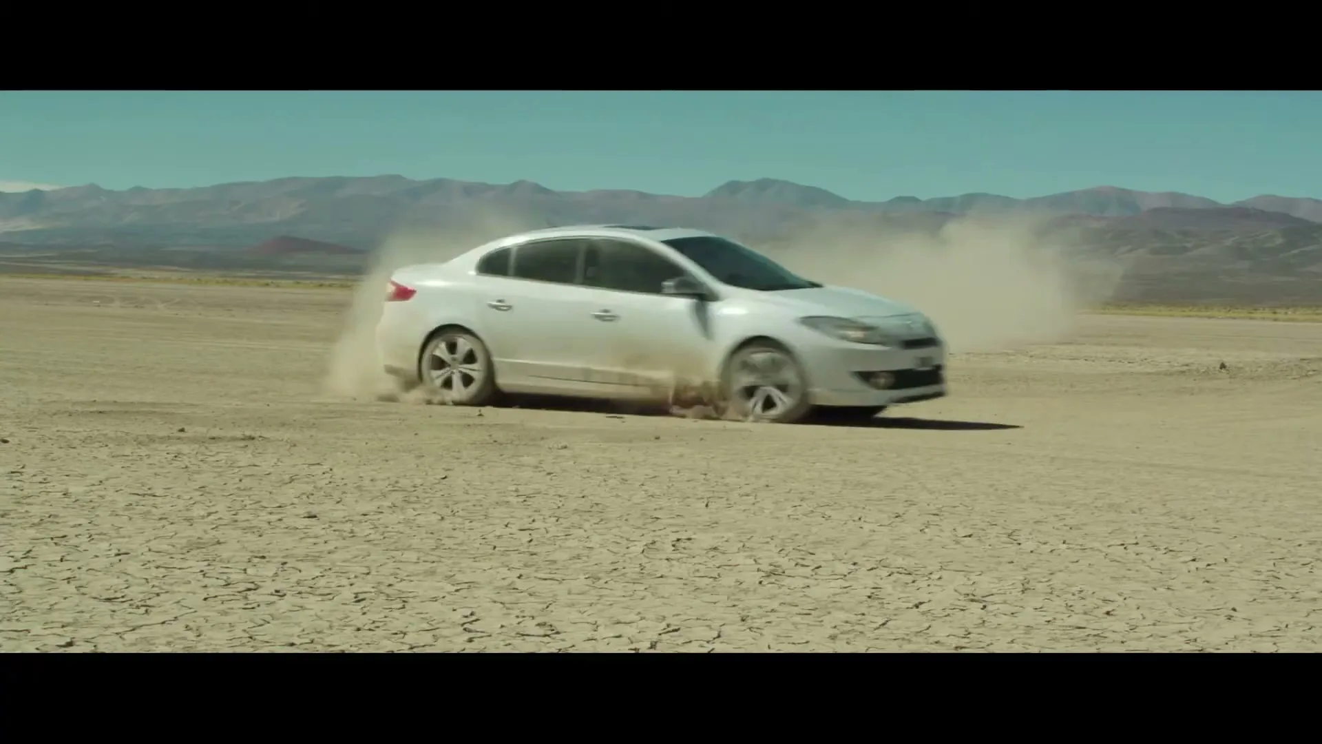 A white sedan drifts on a cracked desert plain, kicking up dust, with mountains in the background under a clear sky.