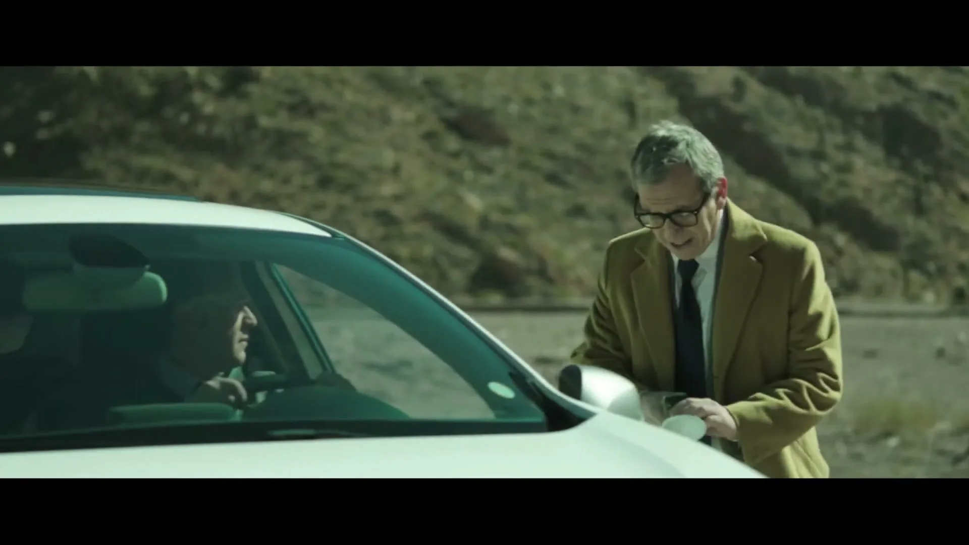 A middle-aged man in a suit and glasses leans into a white car, talking to the driver on a desert road.