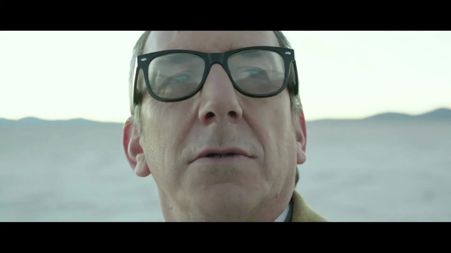 A man in his 50s or 60s, wearing glasses and a brown coat, looks around with a neutral expression in a vast, desolate landscape.