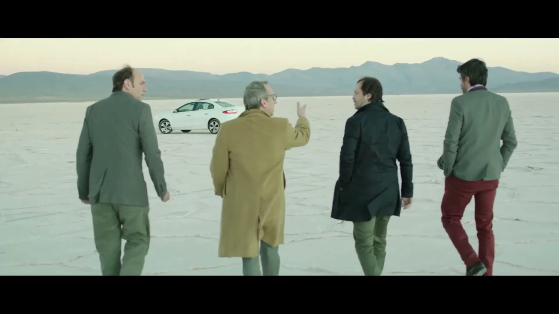 Four men in business casual attire walk away from the camera across a vast, flat, white salt plain towards a white car and distant mountains.