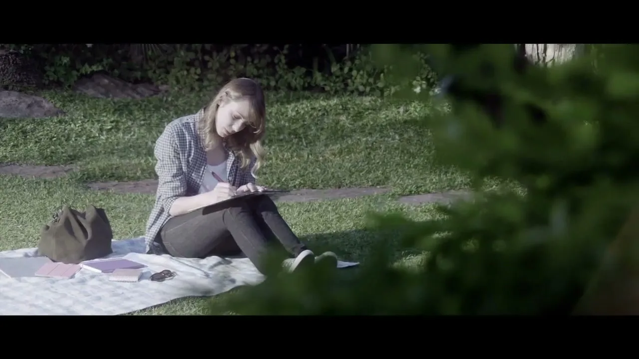 A young woman with blonde hair sits on a blanket in a grassy park, writing in a notebook with a pen.