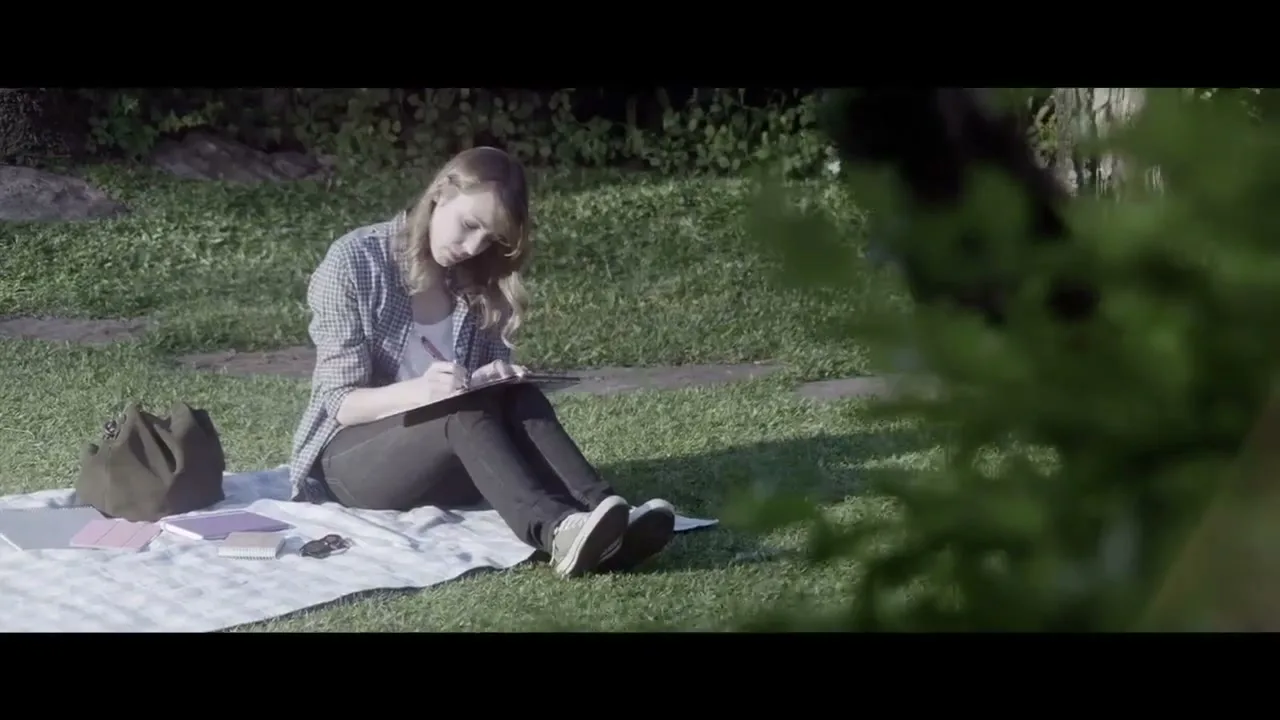 A young woman with blonde hair sits on a blanket in a grassy park, writing in a notebook, with a bag and other items beside her.
