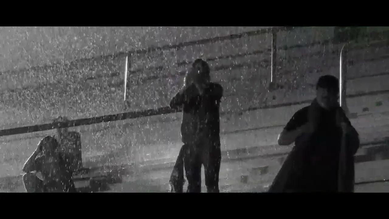 Three men stand on stadium bleachers in the pouring rain at night, two looking up, one looking down, in a black and white shot.