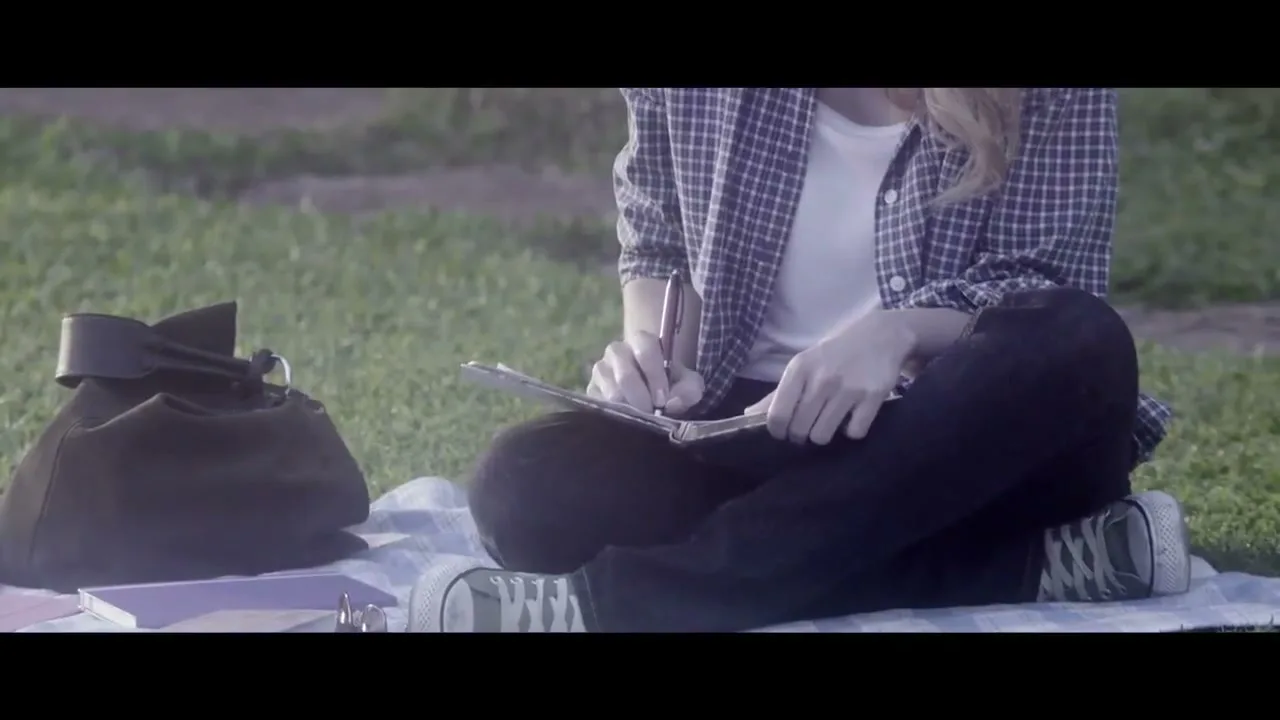 A young woman with blonde hair, wearing a plaid shirt and black pants, sits cross-legged on a blanket in a grassy park, writing in a notebook.