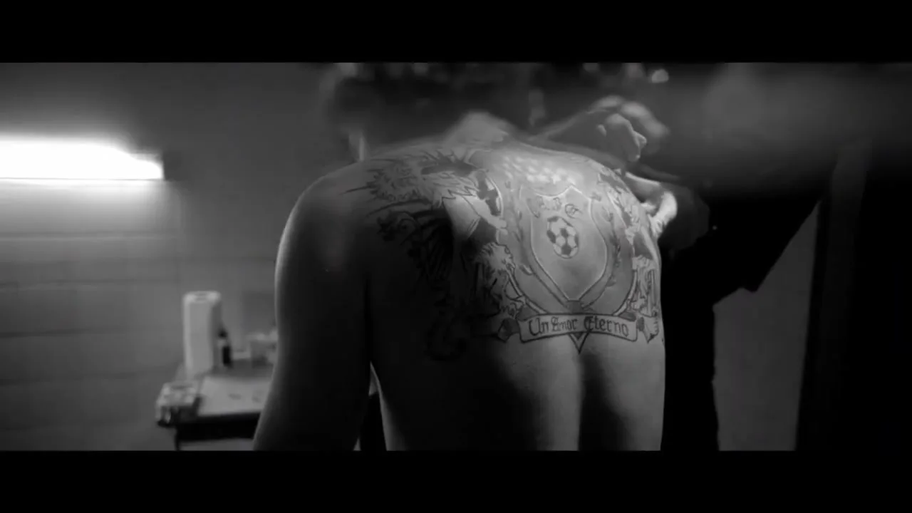A man with a large back tattoo is getting it touched up by a tattoo artist in a dimly lit, industrial-style room.