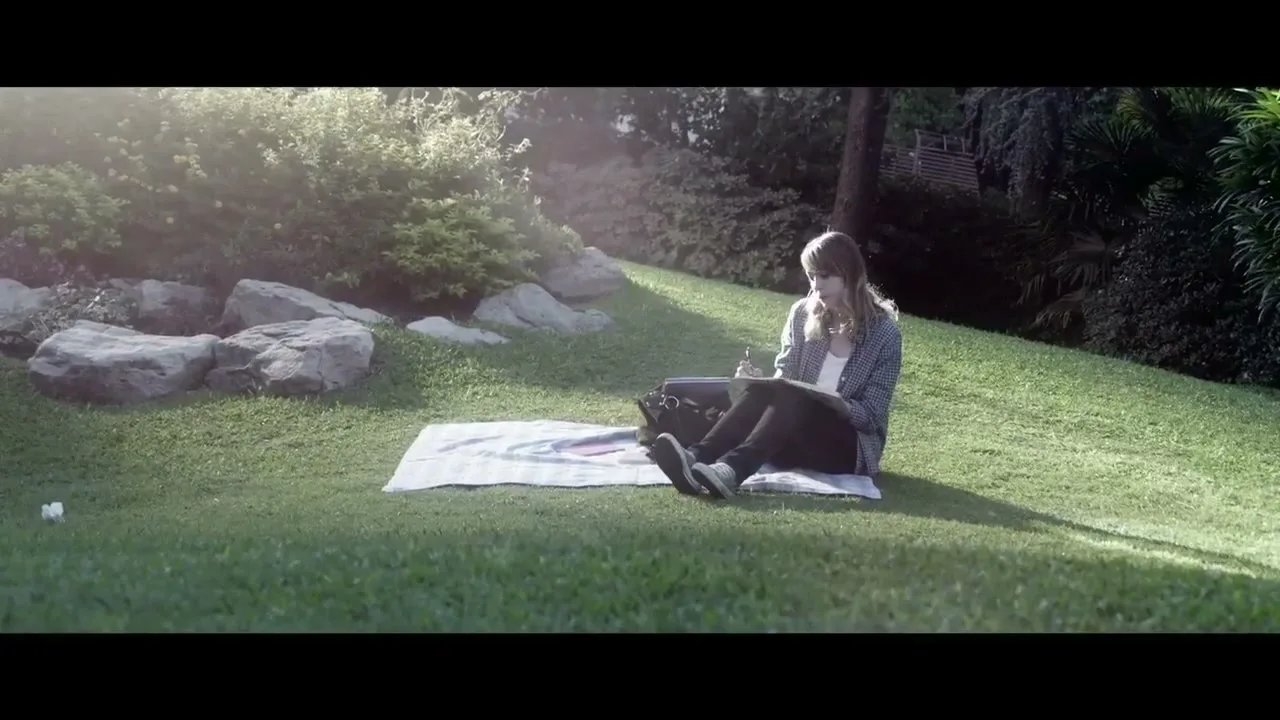 A young woman with long hair sits on a blanket in a sunny park, writing in a notebook.