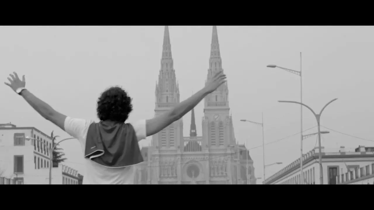A man with curly hair, wearing a white t-shirt and dark pants, kneels on a bridge, then stands and raises his arms in front of a large church.