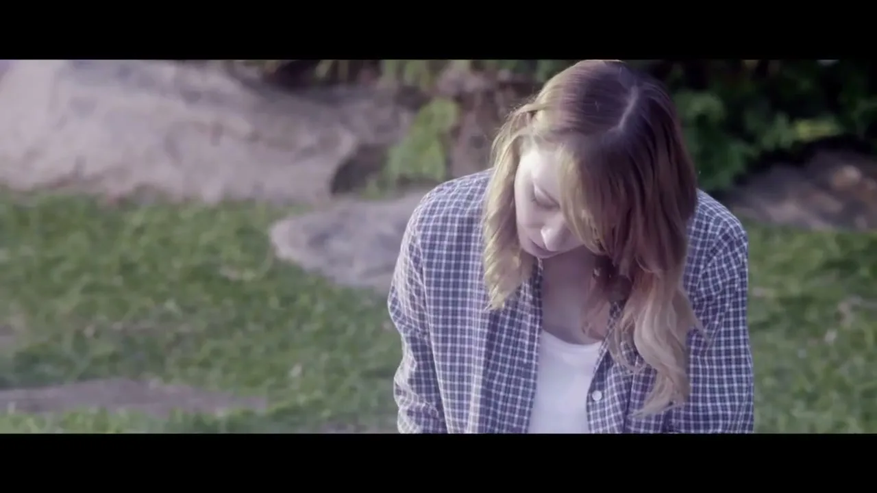 A young woman with blonde hair and a plaid shirt looks down, appearing sad or contemplative, in an outdoor setting with grass and rocks.