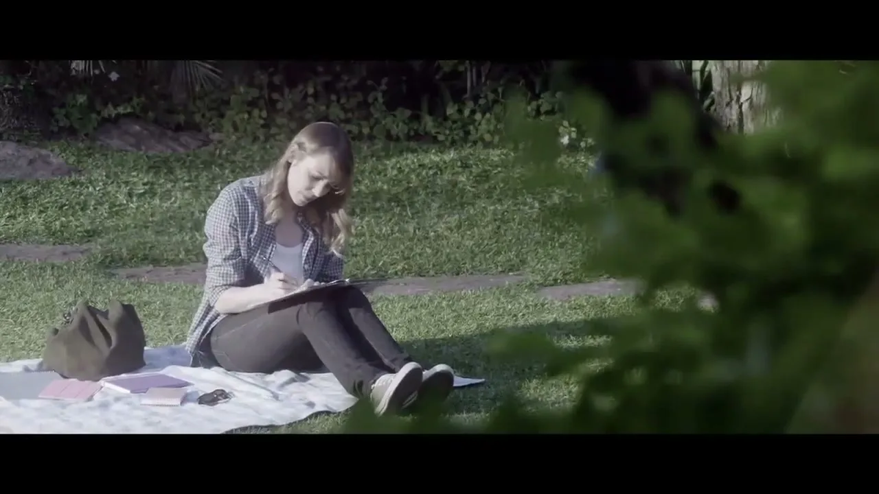 A young woman with long blonde hair, wearing a plaid shirt and black pants, sits on a blanket in a grassy park, writing in a notebook.