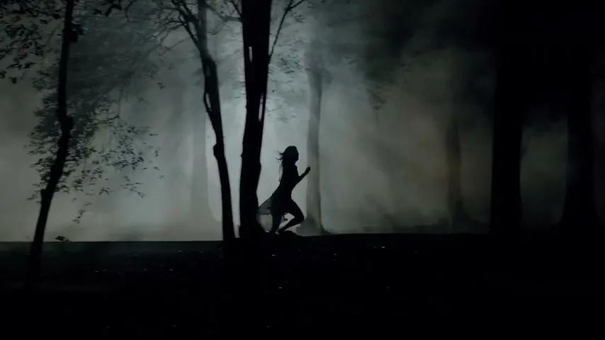 A silhouetted woman with long hair and a flowing dress runs through a dark, misty forest at night, illuminated by shafts of light.
