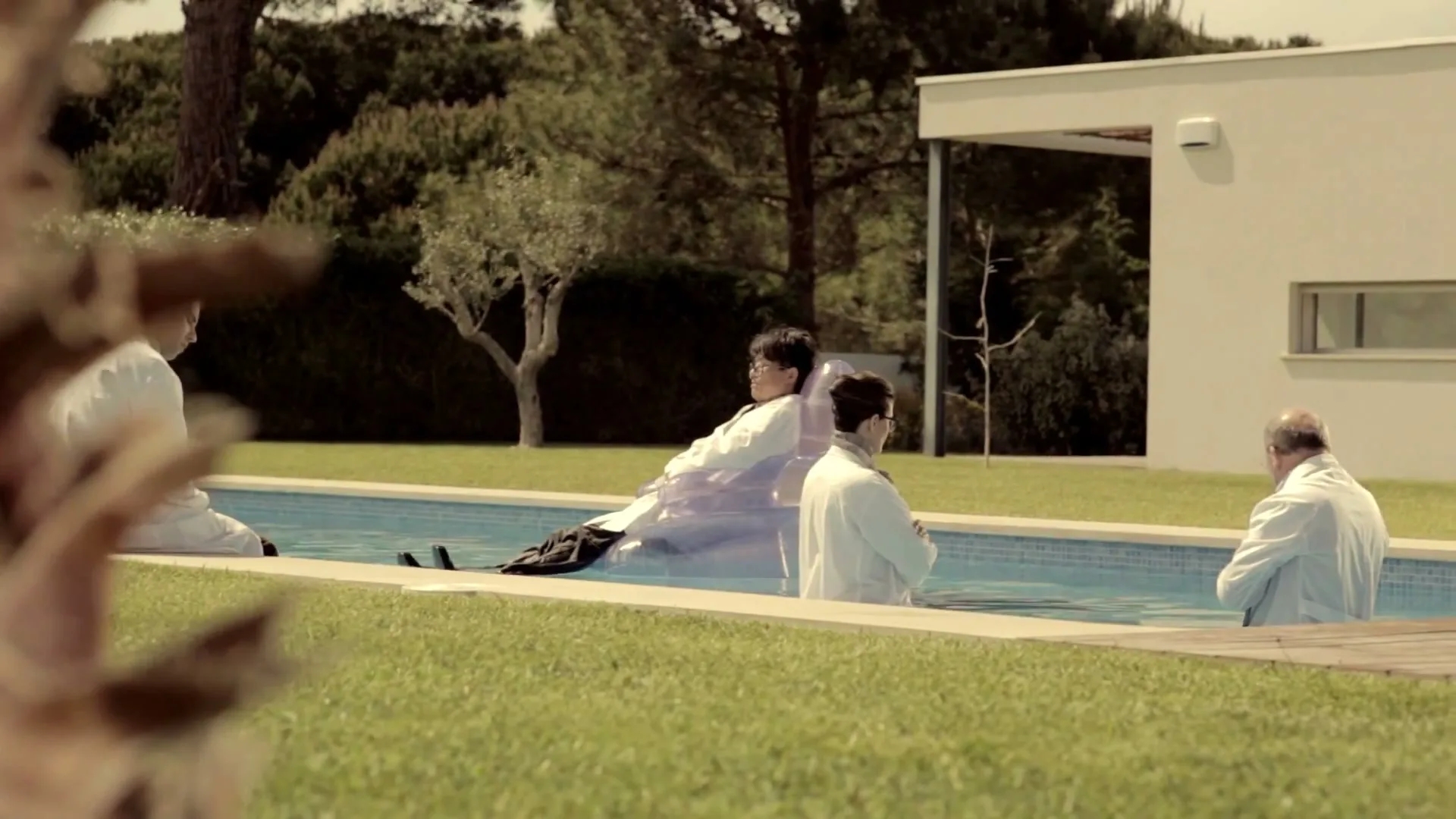 Four scientists in lab coats are in and around a swimming pool in a modern house's backyard on a sunny day.