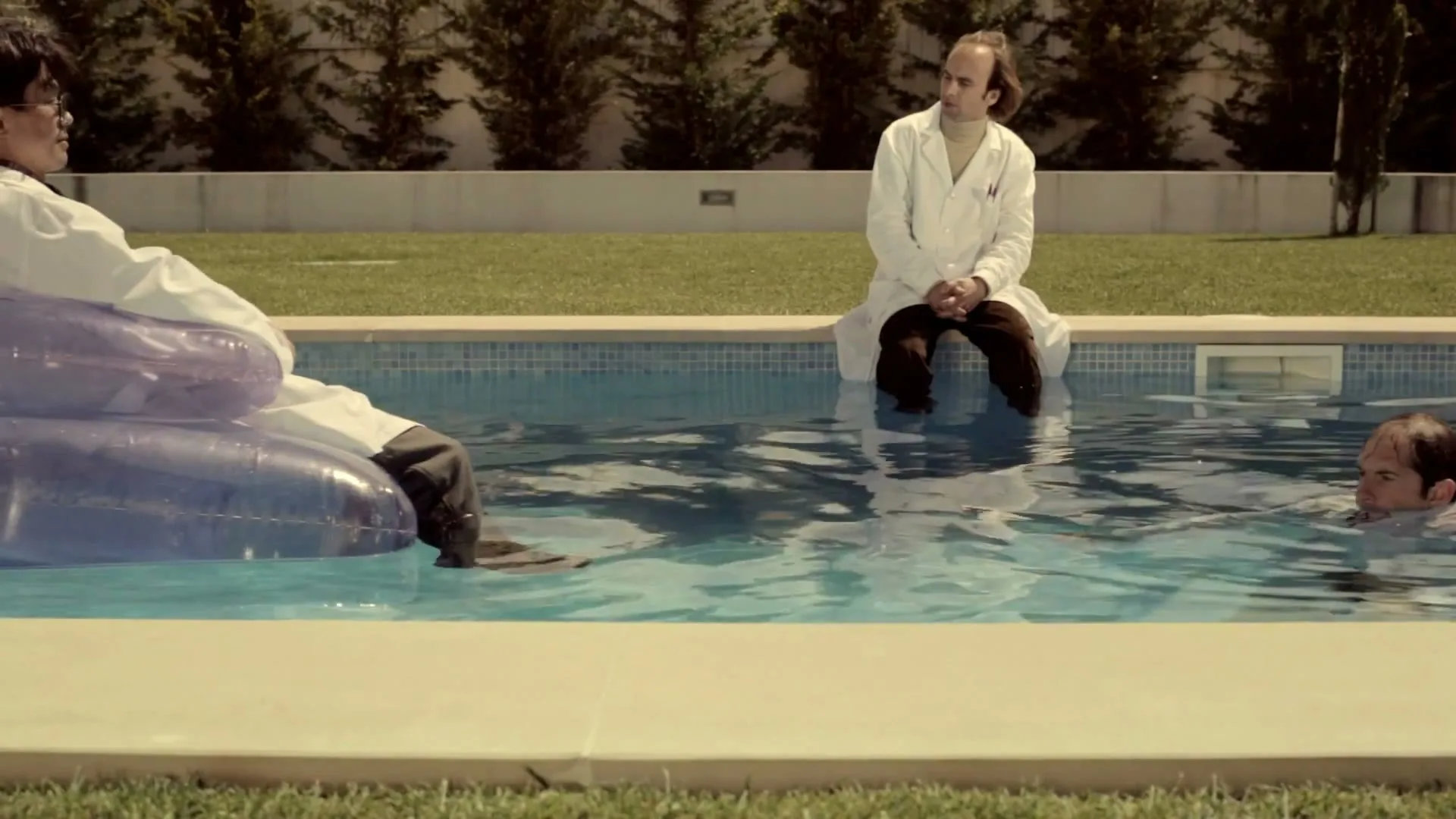 Three male scientists in lab coats are relaxing by a swimming pool, with one on an inflatable, one sitting on the edge, and one in the water.