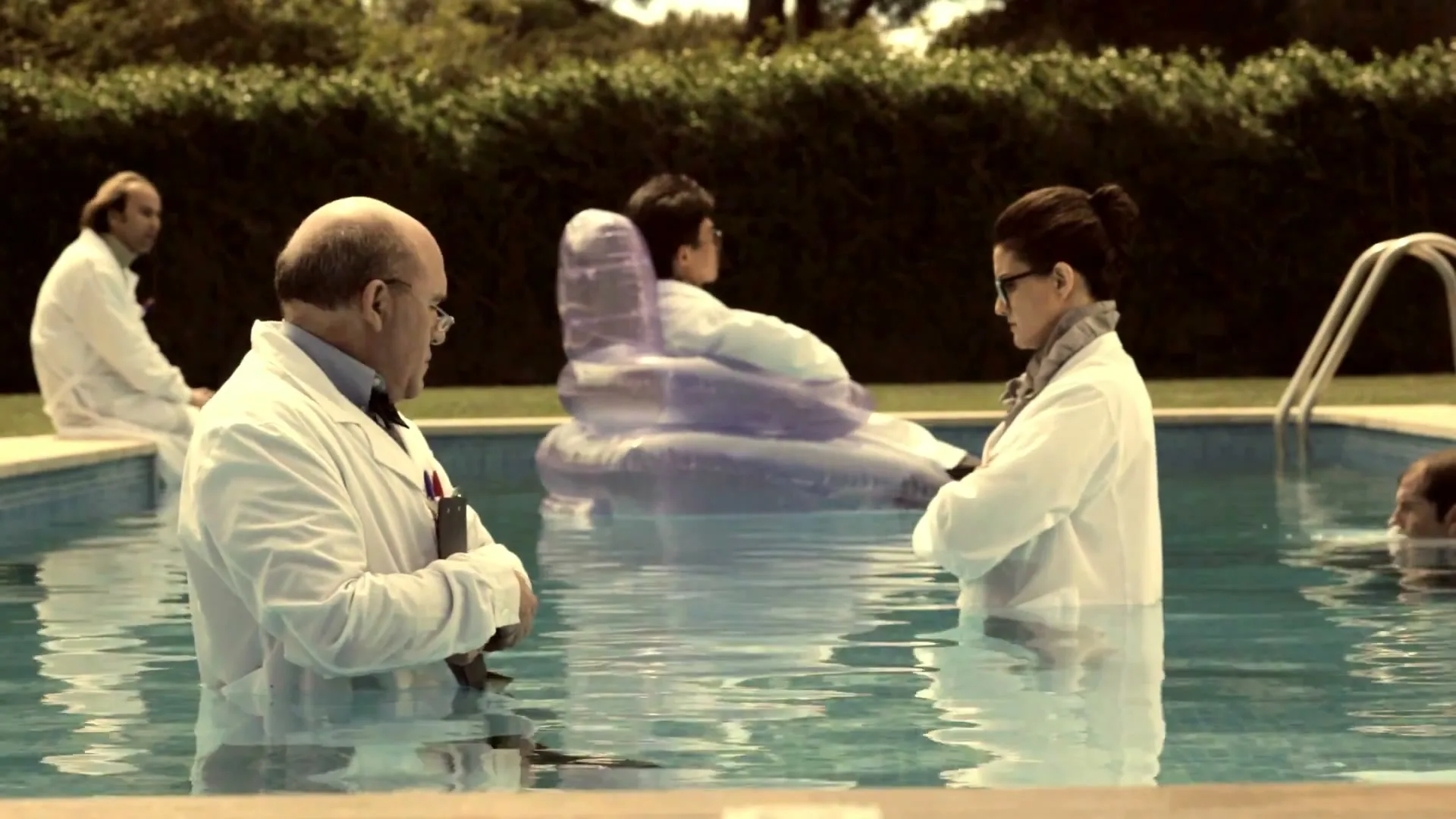A group of scientists in lab coats are standing and floating in a swimming pool, looking at each other in a suburban backyard.
