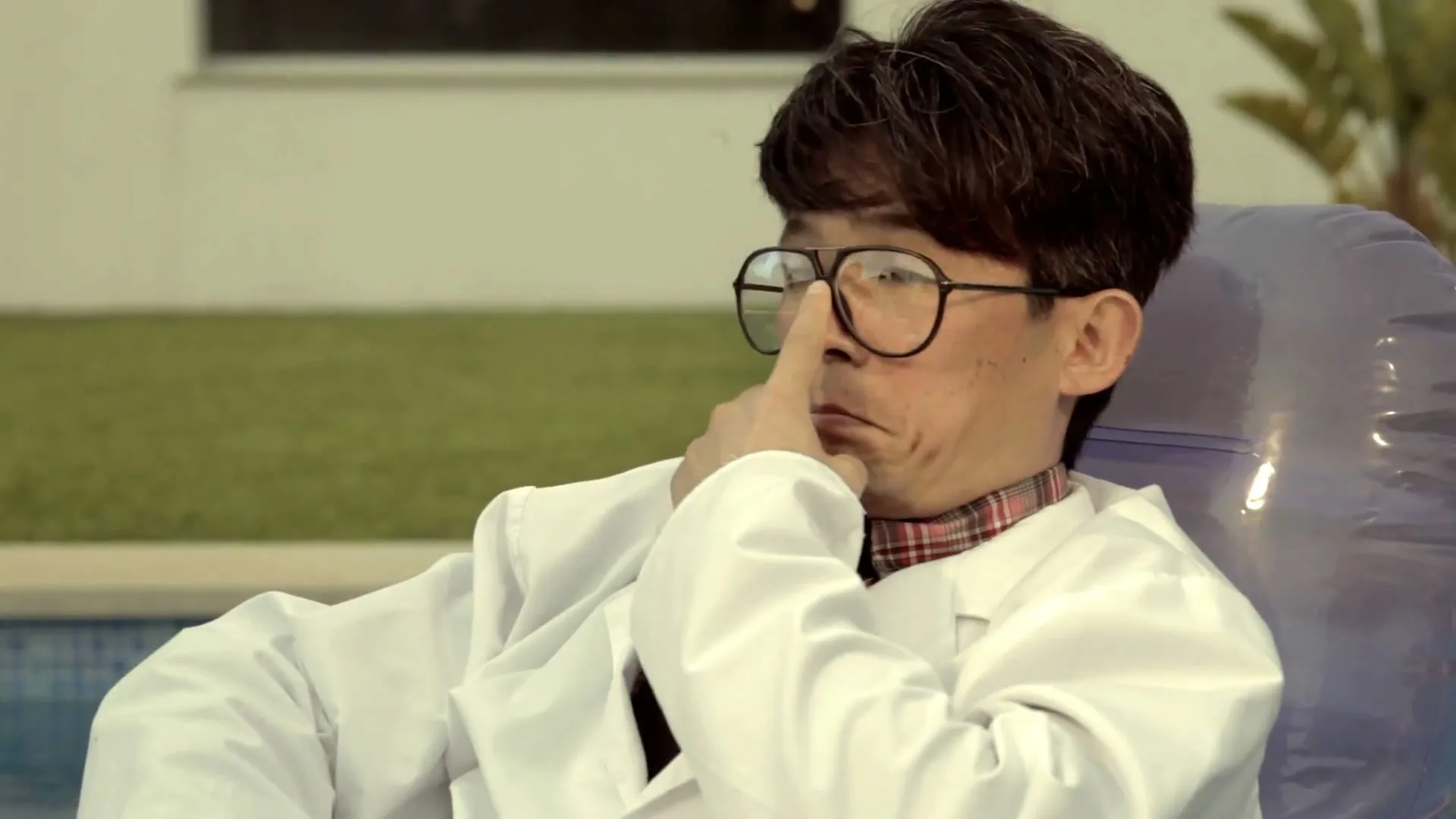A male scientist in a lab coat and glasses sits in an inflatable chair by a pool, adjusting his glasses with a thoughtful expression.
