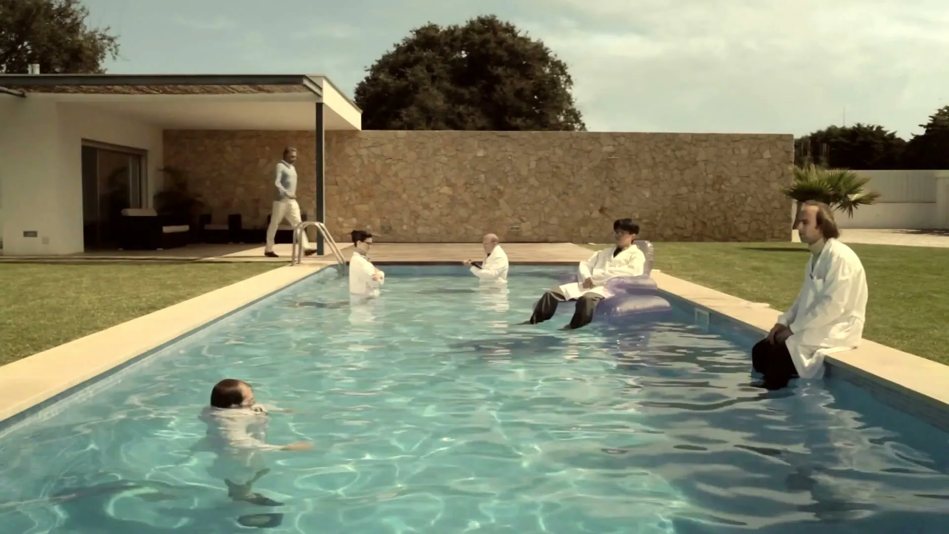 A group of scientists in lab coats are relaxing in and around a swimming pool at a modern house on a sunny day.