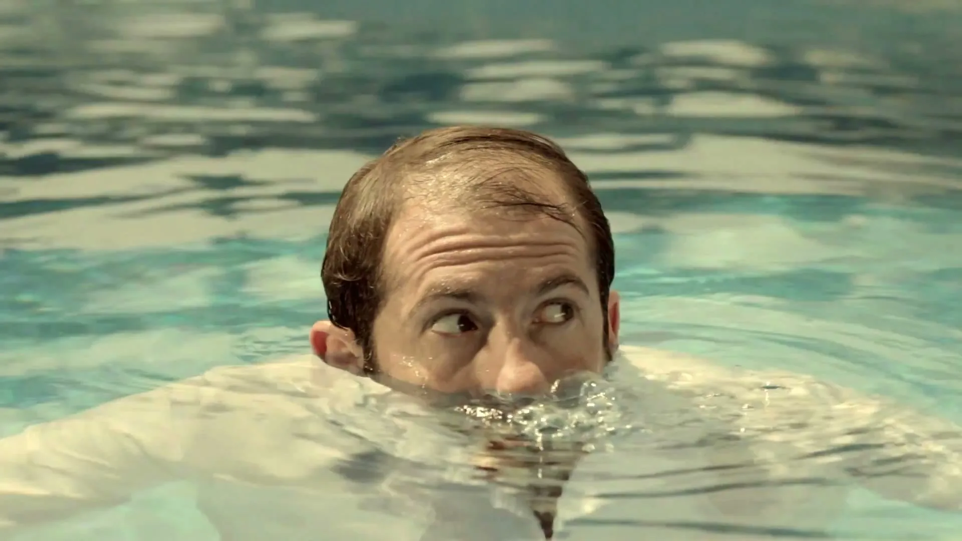A man with wet hair and a white shirt emerges from a swimming pool, looking around with a surprised expression.