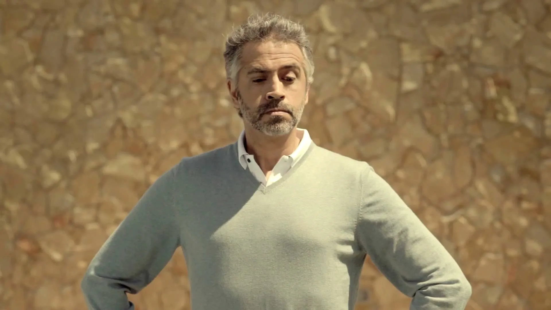 A middle-aged man with grey hair and a beard, wearing a light green sweater, looks down then up with a confused expression against a stone wall.