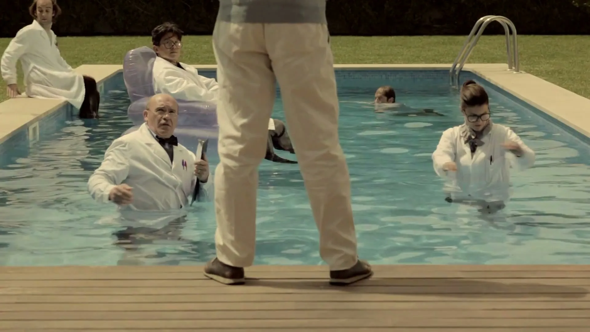 A group of scientists in lab coats are in a swimming pool, looking up at a man standing on the deck, in a suburban backyard.