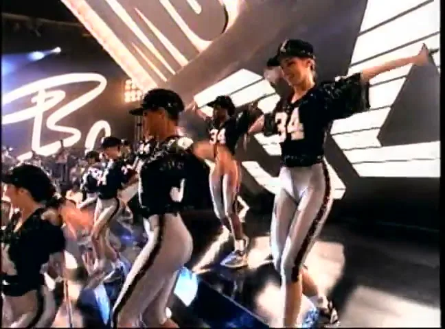 A group of female dancers in football jerseys and caps perform a synchronized routine on a stage with a large "BO" logo in the background.