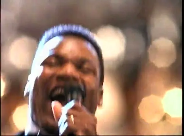 A Black man in his 20s, wearing a tuxedo, sings into a microphone with his eyes closed, on a stage with bokeh lights in the background.