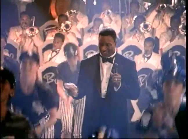 A man in a tuxedo sings into a microphone, surrounded by a brass band and cheering fans in a lively interior setting.