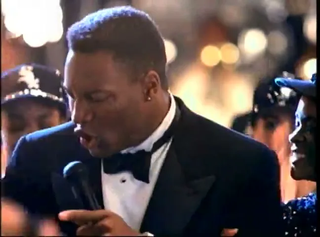 Bo Jackson, a Black man in a tuxedo, sings into a microphone while surrounded by people at a formal event.