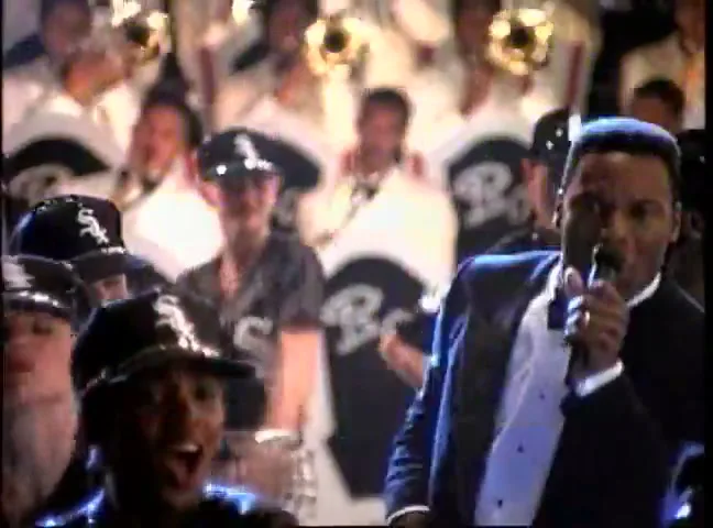 A male singer in a tuxedo performs on stage with a crowd and a brass band behind him, singing into a microphone.