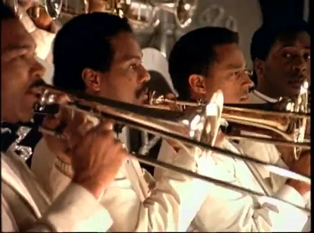 A group of male musicians in white suits play trombones in an orchestra pit, with a focus on their instruments and faces.